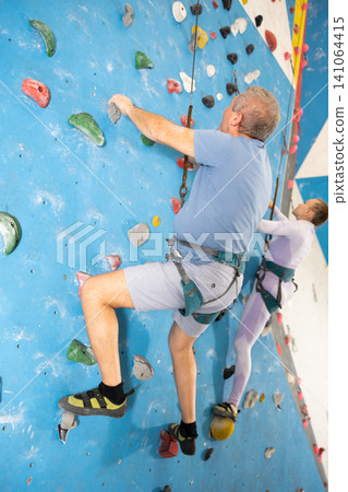 Elderly man in harness with safety rope climbing on bouldering wall Elderly man in harness with safety rope climbing on bouldering wall 141064415