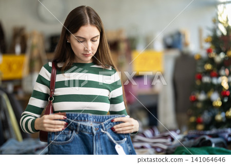 Attentive young woman choosing jeans pantaloons in clothing store Attentive young woman choosing jeans pantaloons in clothing store 141064646