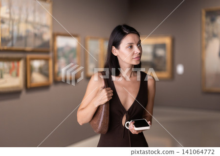 Woman with phone in hands in museum hall, view painting exhibition Woman with phone in hands in museum hall, view painting exhibition 141064772