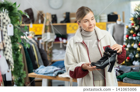 Positive young woman choosing heeled ankle boots in clothing store 141065006