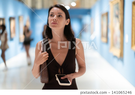 Woman with phone in hands in museum hall, view painting exhibition Woman with phone in hands in museum hall, view painting exhibition 141065246