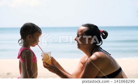 Young mother in swimwear offering a fresh natural lemonade to her little daughter on a tropical beach. Family vacation concept Young mother in swimwear offering a fresh natural lemonade to her little daughter on a tropical beach. Family vacation concept 141068290