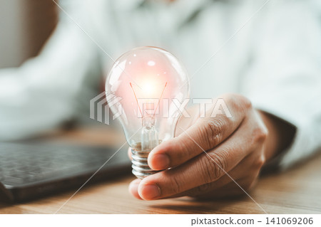 Close up of businessperson holding a glowing light bulb at office desk. Concept of creative thinking, innovative business idea, inspiration, and finding problem solution 141069206
