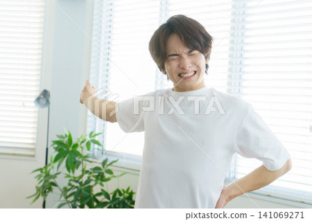 A young Japanese man with a pained expression stretches by the window. A young Japanese man with a pained expression stretches by the window. 141069271
