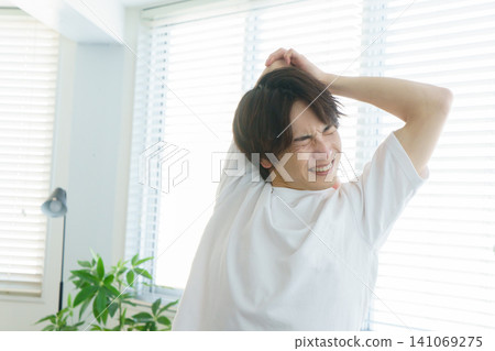 A young Japanese man is stretching by the window, clutching his neck in pain. A young Japanese man is stretching by the window, clutching his neck in pain. 141069275