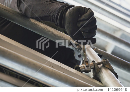 A worker's hand tightening a clamp on scaffolding with a wrench. A worker's hand tightening a clamp on scaffolding with a wrench. 141069648