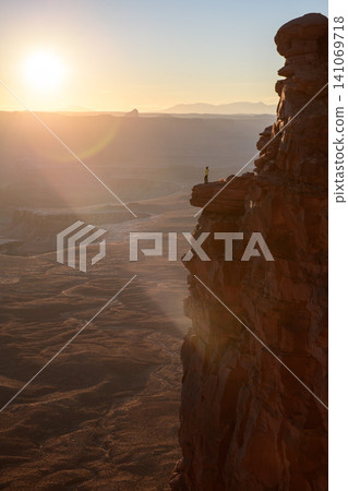 Majestic sunrise illuminating deep canyon of utah while solitary person stands on sheer cliff edge feeling awe and wonder overlooking vast rugged landscape below bathed in golden morning light Majestic sunrise illuminating deep canyon of utah while solitary person stands on sheer cliff edge feeling awe and wonder overlooking vast rugged landscape below bathed in golden morning light 141069718