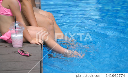 Side close-up view of mother and daughter sitting on pool edge with legs and feet dangling in blue pool water while daughter holds a strawberry cocktail. Summer leisure concept Side close-up view of mother and daughter sitting on pool edge with legs and feet dangling in blue pool water while daughter holds a strawberry cocktail. Summer leisure concept 141069774