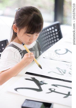A young elementary school girl concentrating on calligraphy, writing characters with a brush; a Japanese child learning calligraphy, with a serious expression; overhead view. A young elementary school girl concentrating on calligraphy, writing characters with a brush; a Japanese child learning calligraphy, with a serious expression; overhead view. 141070210