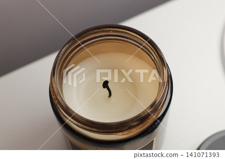 An unlit white wax candle with a black wick rests inside a brown glass jar An unlit white wax candle with a black wick rests inside a brown glass jar 141071393