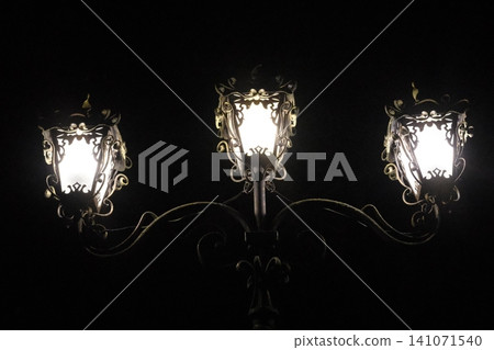 Three antique style lanterns on an ornate streetlamp cast bright light into the deep darkness at night Three antique style lanterns on an ornate streetlamp cast bright light into the deep darkness at night 141071540
