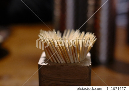 Many light colored toothpicks stand upright in a dark wooden holder on a table indoors Many light colored toothpicks stand upright in a dark wooden holder on a table indoors 141071657