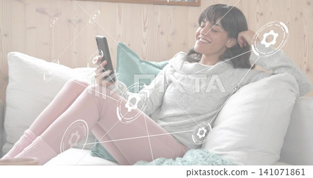 Reclining woman wearing gray sweater, pink leggings scrolling smartphone at home, with pillows 141071861