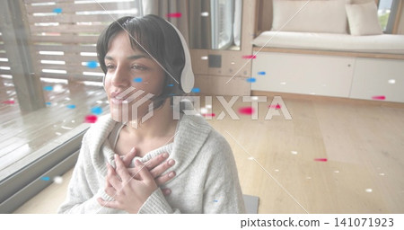 Listening woman with white headset and chunky sweater, sitting on gray mat by sliding glass door 141071923