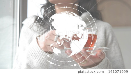 Holding woman in light grey sweater cradling glass teacup and saucer at window, with globe overlay Holding woman in light grey sweater cradling glass teacup and saucer at window, with globe overlay 141071999