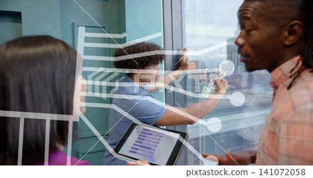 Collaborating trio in plaid, blue, purple tops writing on glass wall in office, tablet showing list Collaborating trio in plaid, blue, purple tops writing on glass wall in office, tablet showing list 141072058
