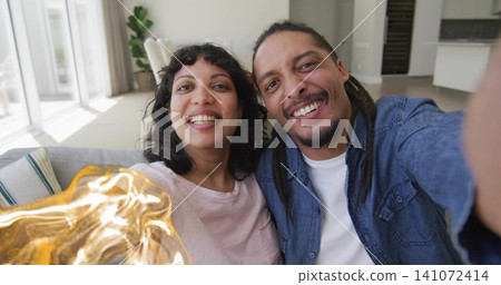 Taking couple wearing light pink T-shirt and blue denim shirt on sofa at home, holding smartphone 141072414