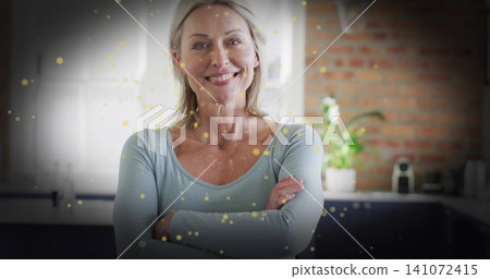 Smiling woman in blue top, thin-chain posing arms crossed in home kitchen, gold bokeh, coffee maker Smiling woman in blue top, thin-chain posing arms crossed in home kitchen, gold bokeh, coffee maker 141072415
