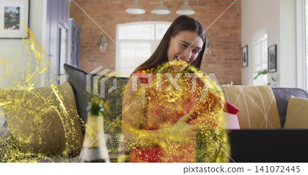 Holding red gift, woman in red top smiling in living room with laptop, champagne and glitter Holding red gift, woman in red top smiling in living room with laptop, champagne and glitter 141072445