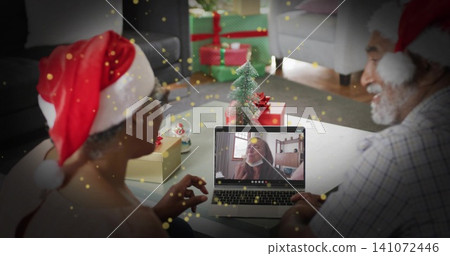 Smiling couple wearing red-and-white Santa hats waving at laptop on living room table, with gifts Smiling couple wearing red-and-white Santa hats waving at laptop on living room table, with gifts 141072446