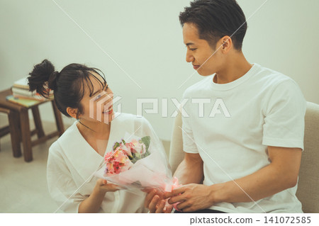 A sofa scene featuring a couple holding a bouquet of flowers and gazing at each other with smiles on their faces. A sofa scene featuring a couple holding a bouquet of flowers and gazing at each other with smiles on their faces. 141072585