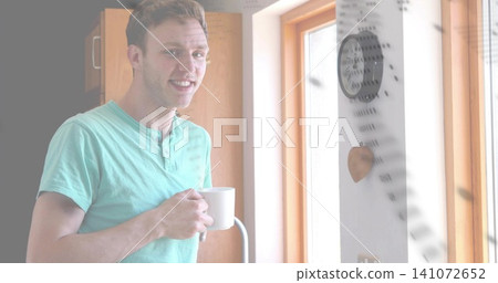 Smiling man in mint-green henley shirt standing in kitchen corner, holding white ceramic mug Smiling man in mint-green henley shirt standing in kitchen corner, holding white ceramic mug 141072652