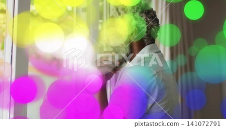 Standing man holding chin, exhaling vapor in room near window, wearing light shirt, colored bokeh Standing man holding chin, exhaling vapor in room near window, wearing light shirt, colored bokeh 141072791