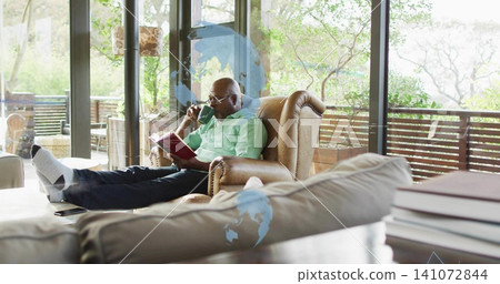 Reading senior wearing green shirt lounging in tan leather armchair in living room holding red book Reading senior wearing green shirt lounging in tan leather armchair in living room holding red book 141072844