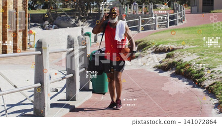 Walking senior man wearing red tank top and black shorts on seaside path, holding orange basketball Walking senior man wearing red tank top and black shorts on seaside path, holding orange basketball 141072864