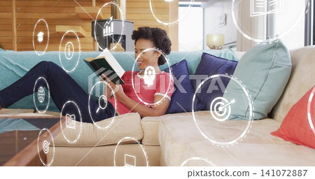 Reclining woman reading hardcover book on living room sofa with digital icons, wearing red top Reclining woman reading hardcover book on living room sofa with digital icons, wearing red top 141072887