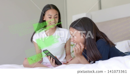 Watching sisters wearing white tee, navy top, sharing smartphone in bedroom at home, code overlay 141073361