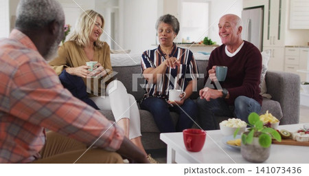 Talking group of four adults sitting around white coffee table in living room, holding ceramic mugs Talking group of four adults sitting around white coffee table in living room, holding ceramic mugs 141073436