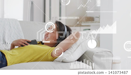 Reclining Korean woman wearing yellow top and grey headphones, holding phone on sofa with AR HUD 141073487