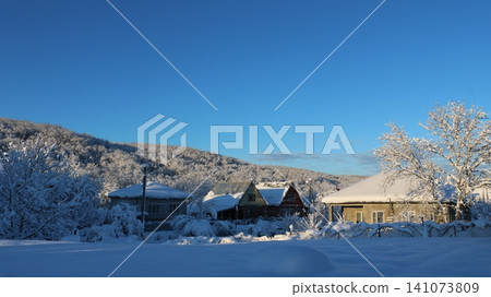 A clear, frosty day in a rural village. Houses with roofs covered in a thick layer of snow stand at the foot of a forested hill under a clear, bright blue sky, with a blanket of snow in the foreground A clear, frosty day in a rural village. Houses with roofs covered in a thick layer of snow stand at the foot of a forested hill under a clear, bright blue sky, with a blanket of snow in the foreground 141073809