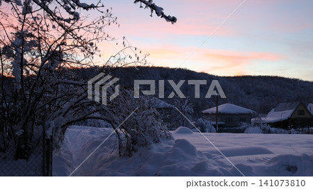 A snow-covered rural landscape. In the foreground are bushes and a path covered in thick snow, while in the distance, white-roofed houses and a wooded hill are visible against a soft pink-blue sky. A snow-covered rural landscape. In the foreground are bushes and a path covered in thick snow, while in the distance, white-roofed houses and a wooded hill are visible against a soft pink-blue sky. 141073810