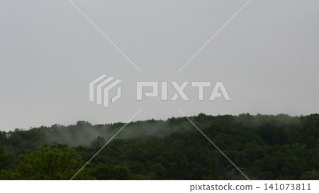 An overcast natural landscape with a dense green forest on the hills. A light white mist swirls above the treetops, and the upper half of the frame is occupied by a gloomy sky covered with dense sky An overcast natural landscape with a dense green forest on the hills. A light white mist swirls above the treetops, and the upper half of the frame is occupied by a gloomy sky covered with dense sky 141073811
