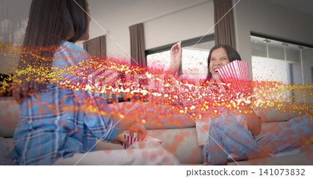 Laughing adult woman and teenage girl sharing popcorn on sofa at home, wearing pajama loungewear Laughing adult woman and teenage girl sharing popcorn on sofa at home, wearing pajama loungewear 141073832