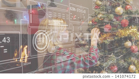 Reaching plaid-shirted boy placing red ornament on Christmas tree at home, HUD panels and fireplace 141074172