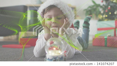 Lying Santa-hat child gazing at snow globe at home on grey carpet, with green overlay Lying Santa-hat child gazing at snow globe at home on grey carpet, with green overlay 141074207