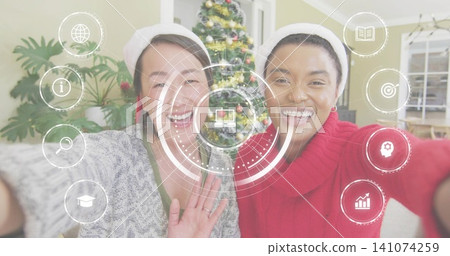 Smiling two women waving and leaning together in home living room, wearing Santa hats, HUD overlay Smiling two women waving and leaning together in home living room, wearing Santa hats, HUD overlay 141074259