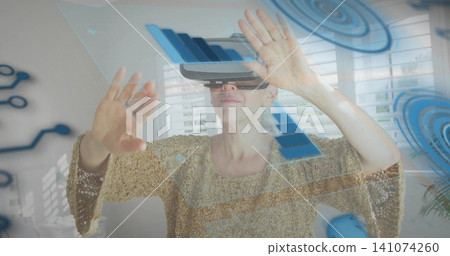 Reaching mature woman wearing VR headset moving holographic charts in living room, beige sweater 141074260