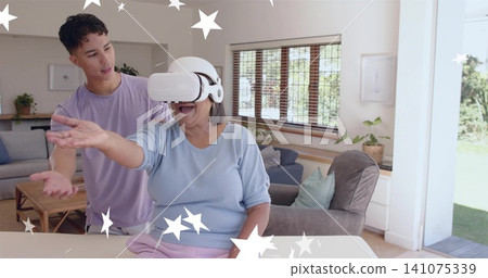 Reaching senior woman with white VR headset, blue top, at home table, man guiding amid star-effects Reaching senior woman with white VR headset, blue top, at home table, man guiding amid star-effects 141075339