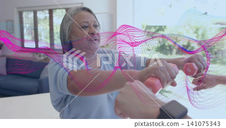 Holding pink dumbbells, senior woman in blue top extending at home counter, helper steadying weight Holding pink dumbbells, senior woman in blue top extending at home counter, helper steadying weight 141075343