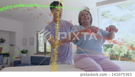 Guiding senior woman in blue top holding pink weights, caregiver supporting on padded table at home Guiding senior woman in blue top holding pink weights, caregiver supporting on padded table at home 141075363
