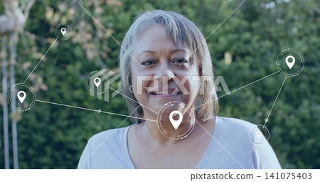 Standing senior woman wearing light gray scoop top and hoop earrings in garden, showing map pins Standing senior woman wearing light gray scoop top and hoop earrings in garden, showing map pins 141075403