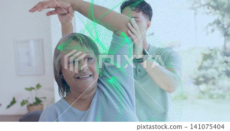 Helping polo caregiver guiding senior in gray top stretching arm in living room, watch, green mesh Helping polo caregiver guiding senior in gray top stretching arm in living room, watch, green mesh 141075404