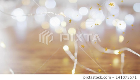 Twinkling thin silver wire fairy lights reflecting on warm wooden tabletop, with gold star confetti Twinkling thin silver wire fairy lights reflecting on warm wooden tabletop, with gold star confetti 141075571