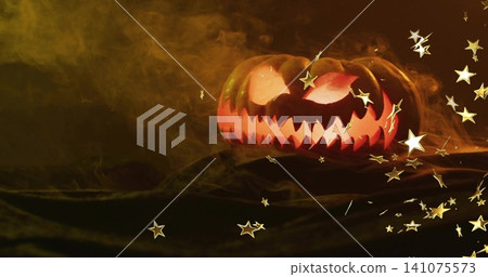 Glowing jack-o'-lantern casting glow in studio on drape with smoke, gold star confetti, copy space Glowing jack-o'-lantern casting glow in studio on drape with smoke, gold star confetti, copy space 141075573