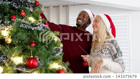 Decorating couple placing red bauble on Xmas tree in living room, wearing Santa hats and sweaters Decorating couple placing red bauble on Xmas tree in living room, wearing Santa hats and sweaters 141075679
