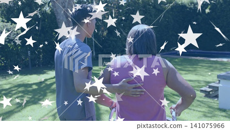 Walking caregiver in gray scrub steadying senior woman in pink top using walker across yard, stars Walking caregiver in gray scrub steadying senior woman in pink top using walker across yard, stars 141075966